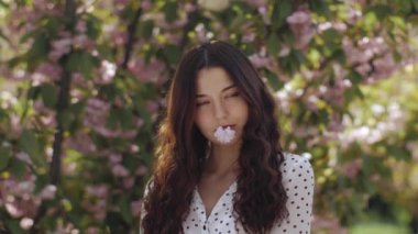 Koyu saçlı, dingin bir genç kadın sakura ağaçlarının altında dikilirken bir çiçeği ağzında tutuyor. Yavaş çekim görselleri bahar zarafetini vurgular ve güzelliği ön plana çıkarır..