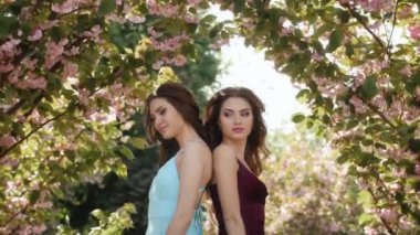 İkiz modeller zarif bir şekilde canlı sakura çiçeklerinin arasında duruyor, şık kıyafetler giyiyorlar. Bahar için ideal, moda editörleri, güzellik kampanyaları ve kardeş sevgisi temaları. 