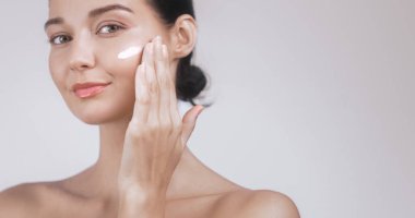 Genç beyaz kadın yanağına yüz kremi sürüyor, cilt bakımını, güzelliği ve kendine bakımı teşvik ediyor. Kozmetik, sağlık ve kişisel bakım ürünü reklamları için ideal.. 