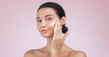Işıl ışıl cildi olan genç ve güzel bir kadın yüzüne krem sürüyor. Pastel pembe arka plana hafifçe gülümsüyor. Bu görüntü güzelliğin ve özgüvenin özünü mükemmel bir şekilde yansıtıyor, ideal.