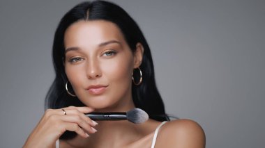 Kendine güvenen bir kadının yüzüne makyaj fırçası tutarken, güzelliği ve cilt bakımını ön plana çıkarırken. Kozmetik, güzellik dersleri ve zarafeti vurgulayan cilt reklamları için ideal.