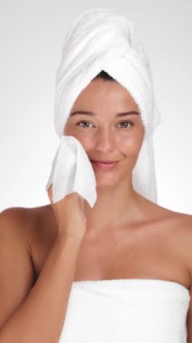 Havluya sarılmış genç bir kadın, parlak, minimalist bir stüdyoda gülümseyen yumuşak bir bezle yüzünü nazikçe temizler. Cilt bakımı, güzellik ve kaplıcalarla ilgili kavramlar için mükemmel.. 
