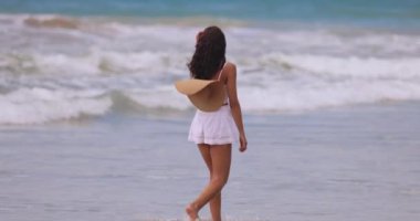 Sri Lanka 'da tropik bir plajda gezinen genç bir kadının sakin bir sahnesi. Beyaz bir elbise giyerek, dalgaların rahatlatıcı sesini, palmiye ağaçlarını ve arka planda bir tatil köyünü seviyor.. 