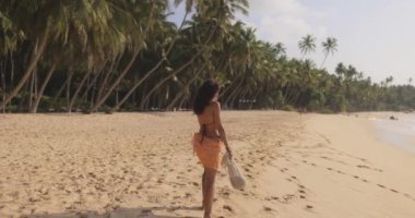 Bikini ve sarong giymiş bir kadın el değmemiş tropikal bir plaj boyunca palmiye ağaçlarıyla kaplı, bir çanta taşıyarak geziniyor. Seyahat, yaz tatili veya gevşemeyi vurgulayan tropikal yaşam tarzı terfileri için ideal