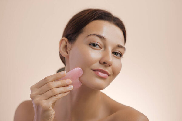 Крупный план молодой белой женщины с сияющей кожей с использованием розового камня гуа ша на лице, способствующей естественной красоте и уходу за собой. Идеально подходит для концепций skincare, spa и wellness.