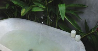 Tropikal bitkiler ve mum ışığında suyla dolu sakin bir taş küvet, rahatlama ve lüks için spa benzeri bir ortam sunuyor. Refah ve kişisel bakım temaları için ideal.