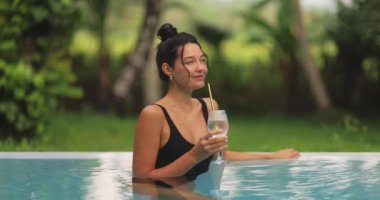 Parlak siyah mayo giymiş güzel bir kadın yemyeşil bir havuzda uzanırken içkisini yudumluyor. Bu lüks sahne rahatlama ve tropikal eğlence temalarını çağrıştırıyor..