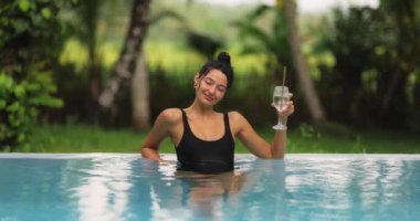 Parlak siyah mayo giymiş neşeli bir kadın tropik bir havuzda dinlenirken ferahlatıcı bir içecek tutuyor. Ağır çekimde yakalanan bu çekim, lüks ve kaygısız bir tatil anını vurgular..