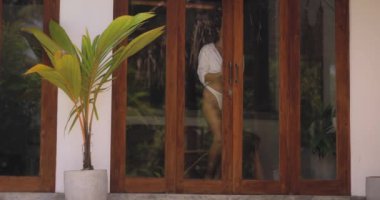 Beyaz tişörtlü, kendine güvenen bir kadın tropikal bir villanın kapısında duruyor ve sıcak havanın tadını çıkarıyor. Seyahat, yaz modası ve yaşam tarzı temaları için ideal.