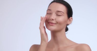 Pırıl pırıl, pürüzsüz bir teni olan genç bir kadın yüzüne nazikçe cilt kremi sürüyor. Minimalist bir stüdyoda gülümsüyor. Güzellik, cilt bakımı ve sağlıkla ilgili kavramlar için mükemmel.. 