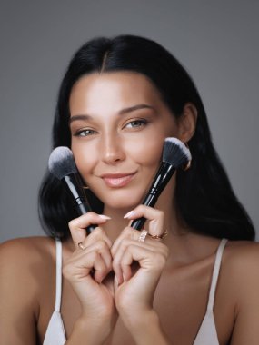 Gülümseyen kadın yüzünün yanında iki makyaj fırçası tutuyor, güzellik aletleri sergiliyor. Kozmetik markalar, makyaj dersleri veya cilt tanıtımları, zarafet ve doğal güzellik için ideal.