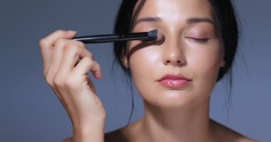 Parlak cildi ve doğal makyajı olan zarif bir kadın fırçayla göz farı sürüyor, yoğun bir odak ve entelektüel. Kozmetik kampanyaları ve güzellik dersleri için mükemmel.. 