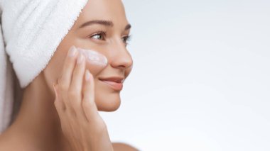 Kafasına havlu sarılmış güzel genç bir kadın gülümserken yanağına yüz kremi sürüyor. Cilt bakımı, güzellik, kişisel bakım reklamları ve promosyonlar için mükemmel.. 