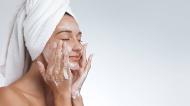 Kafasına havlu sarılmış sakin bir kadın yüzünü köpüklü temizleyiciyle nazikçe temizliyor ve ferahlatıcı bir cilt bakımının keyfini çıkarıyor. Güzelliği, özgüveni ve sağlıklı cildi teşvik etmek için mükemmel.