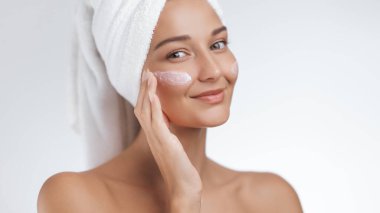 Kafasında havluyla cildini nemlendirmek için yüz kremi süren ve hafifçe gülümseyen genç bir kadın. Güzellik, sağlık, cilt bakımı ve kişisel bakım reklamları için birebir.