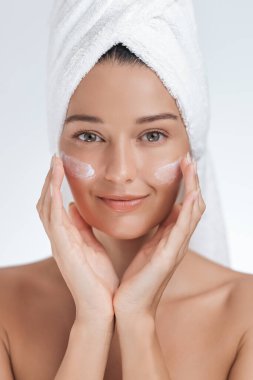Kafasına havlu sarılmış gülümseyen genç bir kadın yanaklarına yüz kremi sürüyor. Cilt bakımı, güzellik ve kişisel bakım reklamları ve sağlık promosyonları için ideal.. 