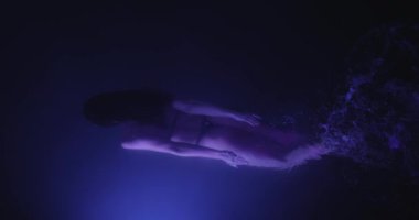 Güzel bir kadın neon ışıklı bir havuzda zarif bir şekilde yüzer. Silueti karanlık sularda baştan çıkarıcı bir şekilde hareket ediyor, mistik mor bir parıltıyla aydınlanıyor, ruhani, rüya gibi bir ışık yaratıyor.