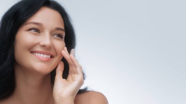 Koyu renk saçlı ve parlak tenli genç bir kadının portresi. Yumuşak beyaz bir arka plana karşı doğal bir şekilde gülümsüyor. Güzellik, cilt bakımı, sağlık, yaşam tarzı kavramları ve promosyonlar için mükemmel..