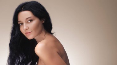 Uzun siyah saçlı zarif bir Asyalı kadın tarafsız bir arkaplanda zarifçe poz veriyor. Güzellik, cilt bakımı ve saç bakım reklamları için ideal. Doğal zarafet ve parlak cilt için..