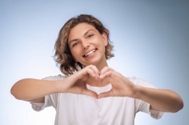 Kısa kıvırcık saçlı neşeli beyaz kadın elleriyle kalp şeklini alıyor ve içten gülümsüyor. Aşk, ilgi, arkadaşlık, mutluluk, sosyal destek ve olumlu duygular kavramı.