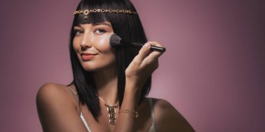 Pürüzsüz, siyah saçlı, Kleopatra 'dan esinlenilmiş altın başlığıyla süslenmiş, makyajını fırçayla yaparken gülümseyen şık bir kadın. Pembeye karşı ışıl ışıl ifadesi ve lüks görünüşü