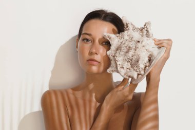 Zarif bir kadın büyük bir deniz kabuğu tutar, doğal ışık ve gölgesi onun sakin ifadesini vurgular. Moda, yaşam tarzı, plaj temalı promosyonlar ve reklamlar için mükemmel..