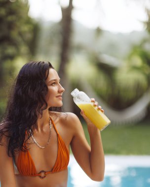 Turuncu bikinili bir kadın, tropik bir ortamda havuzda dinlenirken ferahlatıcı bir şişe meyve suyu yudumluyor. Bu görüntü yazın ve sağlıklı yaşamın özünü yakalar. Sağlıklı yaşam için mükemmeldir.