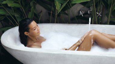 Huzurlu bir kadın, bereketli tropikal bitkilerle çevrili açık hava küvetinde rahatlatıcı bir köpük banyosundan hoşlanır. Huzurlu ortam sağlık, özgüven ve lüks, spa ve yaşam tarzı için mükemmel.