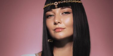 Kendine güvenen, parlak siyah saçlı, Kleopatra 'dan esinlenilmiş altın bir başlıkla süslenmiş pembe bir arka plana hafifçe gülümseyen bir kadın. Işıl ışıl teni ve lüks tarzı güzelliğin temalarını vurguluyor.