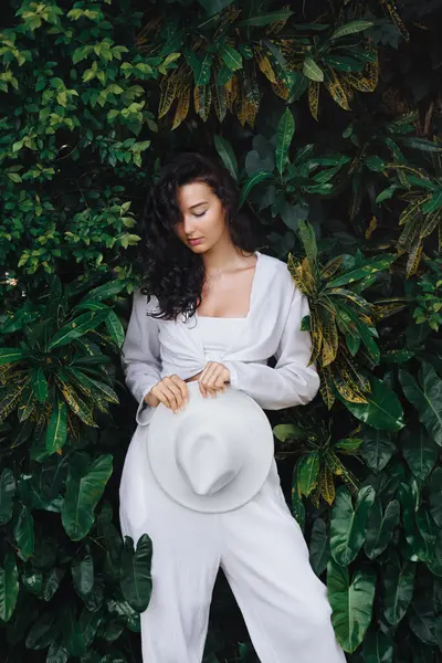 Beyaz bir elbise giymiş güzel esmer kadın zarif bir şekilde beyaz bir şapka tutuyor ve canlı yeşil yaprakların önünde poz veriyor. Moda, doğa ve yaşam tarzı temaları için mükemmel..