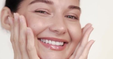 Beyaz kadın parıldayan tenine ağır çekimde masaj yaparken gözleri kapalı gülümser. Cilt bakımı, yüz masajı, kozmetik ürünleri veya güzellik tanıtımı için mükemmel..