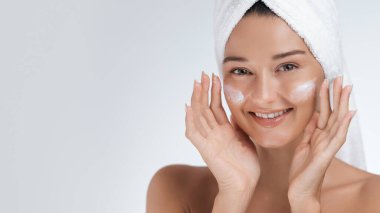 Kafasına havlu sarılmış gülümseyen genç bir kadın yanaklarına yüz kremi sürüyor. Cilt bakımı, güzellik ve kişisel bakım reklamları ve sağlık promosyonları için ideal.. 