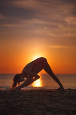 Gün batımında plajda yoga yapan, siluette yakalanmış sağlıklı bir kadın. Esnekliği, farkındalığı ve dengeyi kucaklayarak ileri doğru derin bir dönüş sergiliyor. Sağlık ve meditasyon için mükemmel.