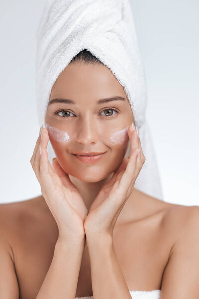 Крупным планом молодая женщина наносит увлажняющий крем на лицо, завернутый в полотенце. Перфект для продвижения красоты, skincare и велнес-продуктов, которые фокусируются на гидратации, самоуходе и перчатках