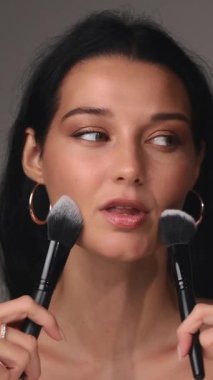 Koyu renk saçlı ve parlak tenli neşeli bir kadın gülümserken iki makyaj fırçası tutuyor. Kendine güven ve güzellik yayarak bu görüntüyü cilt bakımı, kozmetik ve kendine bakım için ideal hale getiriyor.