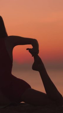 Bir kadın, gün batımında bir plajda, parlayan gökyüzüne ve sakin okyanusa karşı, yoga pozu verir. Sağlığı, esnekliği ve barışçıl zindelik kavramlarını desteklemek için idealdir. Dikey video.