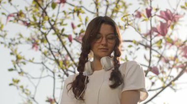 Saç örgülü, retro gözlüklü ve kulaklıklı genç bir kadın güneşli bahar bahçesinde pembe çiçeklerin arasında sessizce duruyor. Gençlik, farkındalık, yaşam tarzı ya da mevsimsel hikaye anlatma görselleri için harika.