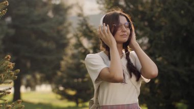 Saçları örgülü, kulaklık takan, güneş gözlüklü, parkta müzik dinlemekten hoşlanan genç bir kadın. Açık hava rahatlama ve müzik zevki konsepti. Sinema yavaş çekim 4k görüntü, reklam için tasarım ve