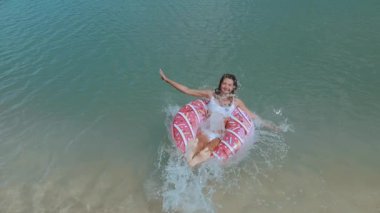 Beyaz bikinili bir kadının tropikal sulara atladığı, pembe çörek şeklindeki şişirilebilir bir çöreğin üzerine mükemmel bir şekilde indiği eğlenceli süper yavaş çekim videosu. Seyahat temaları için canlı bir yaz sahnesi. 