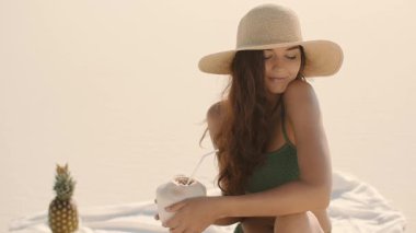 Güneş şapkalı ve yeşil bikinili güzel bir kadın kumlu bir sahilde ağır çekimde otururken taze hindistan cevizinin tadını çıkarıyor. Tatil, tropikal ve yaşam tarzı kavramları için mükemmel..