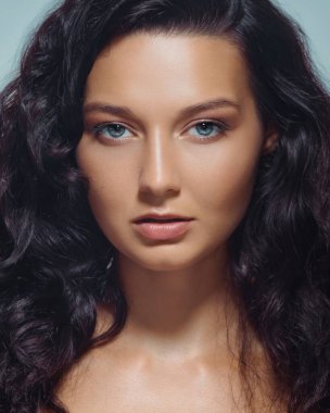 Stüdyoda parlak tenli, açık mavi gözlü ve uzun siyah dalgalı saçlı genç bir kadının yakın çekimi. Cilt bakım ürünleri, kozmetik ürünleri ve moda markaları için çok kaliteli bir görüntü..
