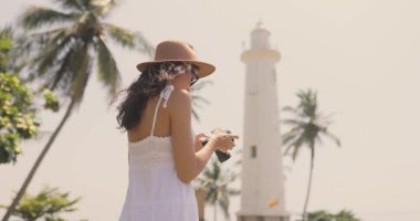 Beyaz yazlık elbiseli bir kadın turist Sri Lanka 'daki Galle Fort deniz fenerinin fotoğraflarını çekiyor. Palmiye ağaçları ve güneşli gökyüzüyle çevrili bu görüntü, sakin ve tropik bir yolculuk anını yakalar.