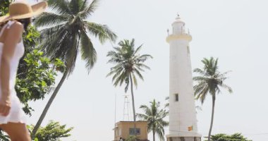 Beyaz elbiseli ve güneş şapkalı bir kadın Sri Lanka 'daki Galle Fort deniz fenerine bakıyor. Tropikal palmiye ağaçları ve parlak gökyüzü ile çevrili bu dingin seyahat sahnesi turizm, tatil ve