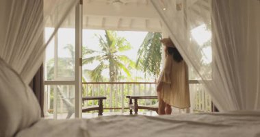 Tropikal bir tatil köyünün balkonunda akmakta olan perdelerle çerçevelenmiş bir kadının sakin görüntüsü. Cennet ortamlarında lüks seyahat, rahatlama ve egzotik tatil temaları için ideal..