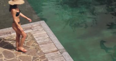 Siyah bikini ve hasır şapkalı şık bir kadın ayaklarını suya daldırmak için oturmadan önce havuz kenarında tropik bir güvertede yürüyor. Yaz, seyahat ve lüks yaşam tarzı kavramları için ideal..