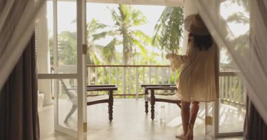 Bir kadın zarif bir şekilde tropikal bir balkonda dikiliyor, perdelerle çerçevelenmiş, yemyeşil manzaranın tadını çıkarıyor. Lüks yaşam tarzı, seyahat promosyonları ve egzotik tatil kavramları için mükemmel..