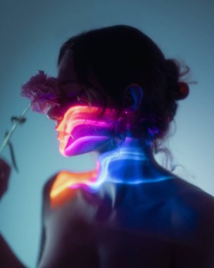 Elinde çiçek tutan genç bir kadının sanatsal silueti, parlak neon ışık projeksiyonlarıyla aydınlatılmış. Modern reklam ve dijital sanat eserleri için kavramsal ve duygusal stüdyo portresi.