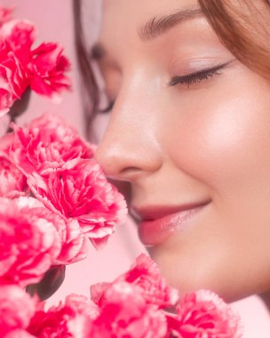Gözleri kapalı gülümseyen, canlı pembe karanfillerin kokusundan zevk alan beyaz bir kadının yakın çekimi. Güzellik kavramı, doğa, tazelik ve çiçekli cilt tanıtımı.