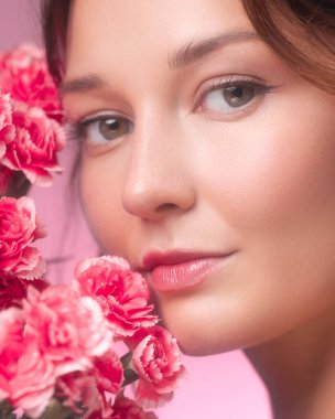 Parlayan derisi ve doğal makyajı olan pembe karanfillerden usulca bakan genç bir kadının güzel yakın çekim stüdyosu. Pastel pembe arka plan ve narin çiçek aranjmanı...