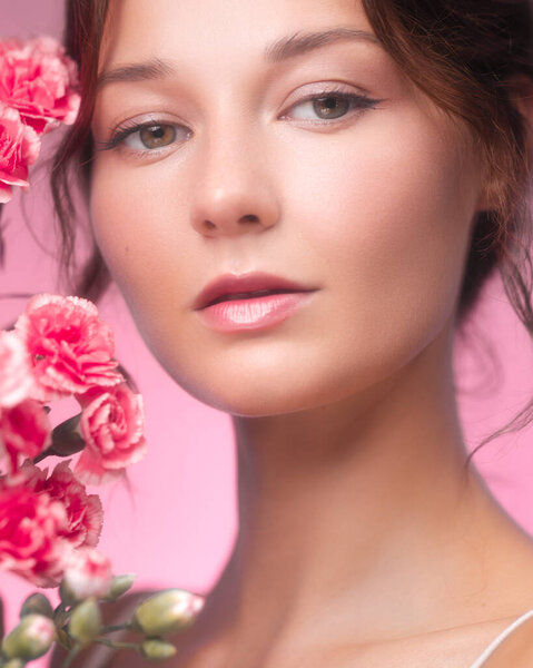 Intimate close-up studio portrait of a stunning young woman with flawless skin and natural makeup, surrounded by vibrant pink carnations. Her calm expression and the soft pink background create a